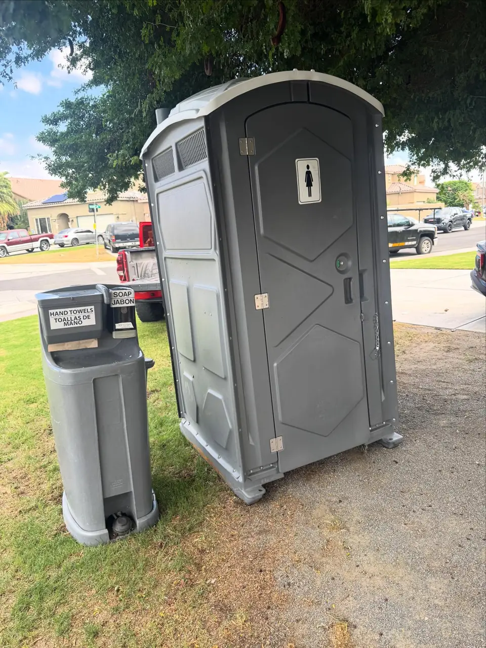 Gray portable toilet on a lawn with a freestanding hand wash station beside it