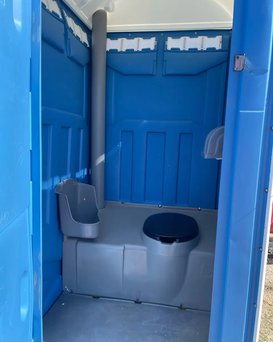 Inside a standard blue porta potty showing the toilet seat, urinal, vent pipe and paper holder
