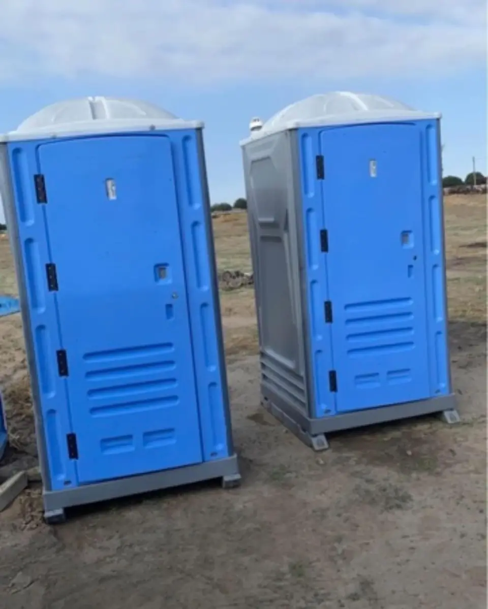 Two blue standard porta potties on a dirt construction lot under a clear sky