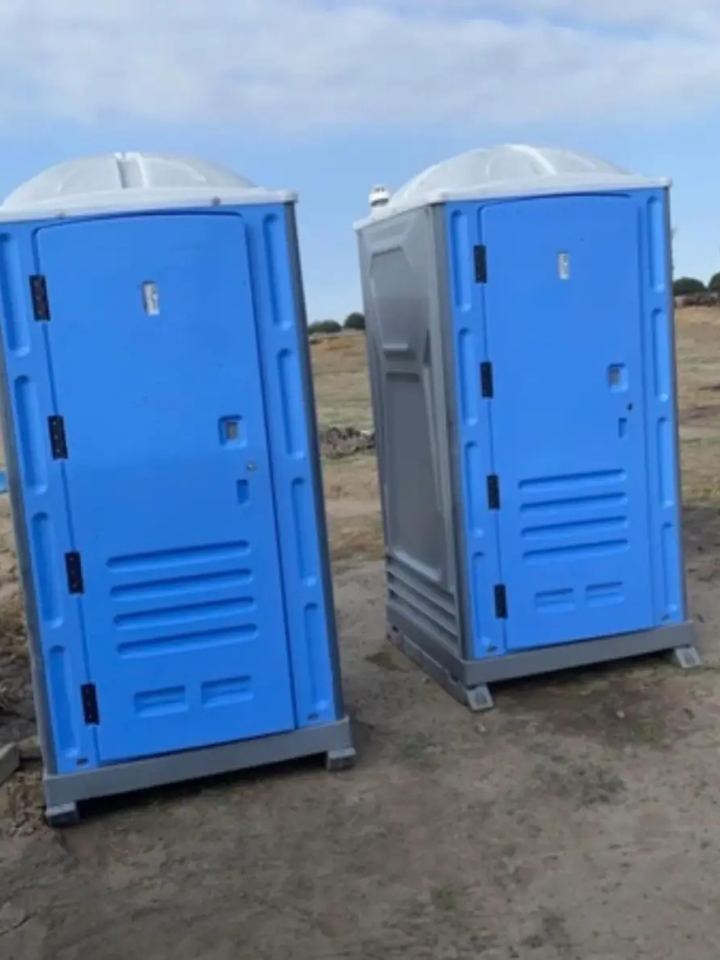 Two blue standard porta potties on a dirt construction lot under a clear sky